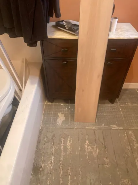 Bathroom during rebuild: old flooring removed down to the subfloor, new plank flooring staged