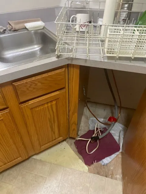 Under a kitchen sink before cleanup: cabinet base removed, supply lines and a towel on the exposed floor